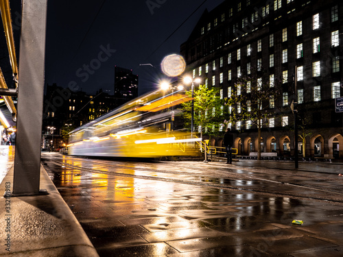 A dynamic night scene showing a Metrolink tram captured with a slow shutter outside St Peter's Square in Manchester, UK