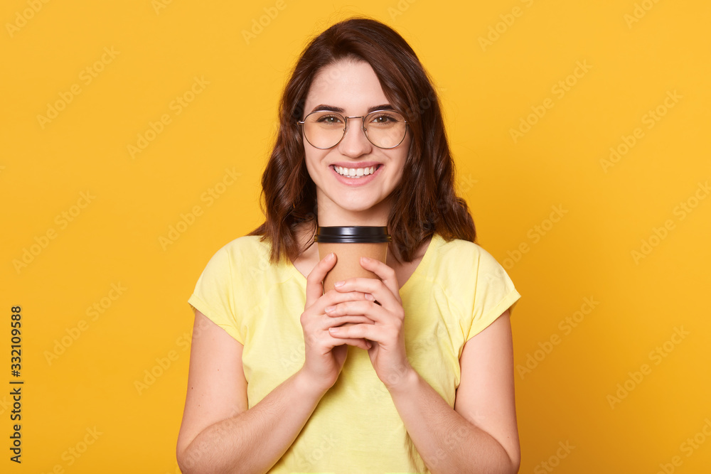 Portrait of smiling happy woman, wearing yellow casual t shirt and glasses, has dark hair, holding paper cup of takeaway coffee, looks directly at camera, isolated overyellow studio background. Portrait of smiling happy woman, wearing yellow casual t shirt and glasses, has dark hair, holding paper cup of takeaway coffee, looks directly at camera, isolated overyellow studio background.