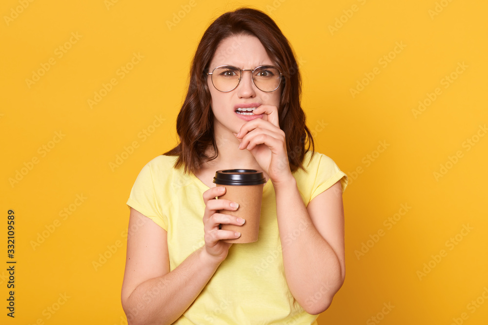 Discontent Caucasian young woman with dark hair frowning her face, having displeased facial expression, holding takeaway coffee in hands while biting her finger, looks sad, dressed casual clothing. Discontent Caucasian young woman with dark hair frowning her face, having displeased facial expression, holding takeaway coffee in hands while biting her finger, looks sad, dressed casual clothing.