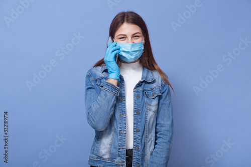 Horizontal shot of young Coucasian woman wearing blue flumask, blue mask, white shirt and denim jacket, female trying to protect from coronavirus, talking to phone with friend. Covid 19 concept. photo