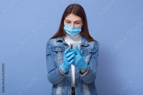 Close up portrait of young brunette woman wearing medical face mask and disposable blue gloves, typing on mobile phone, girl posing isolated over blue background. Coronavirus, covid 19 concept. photo