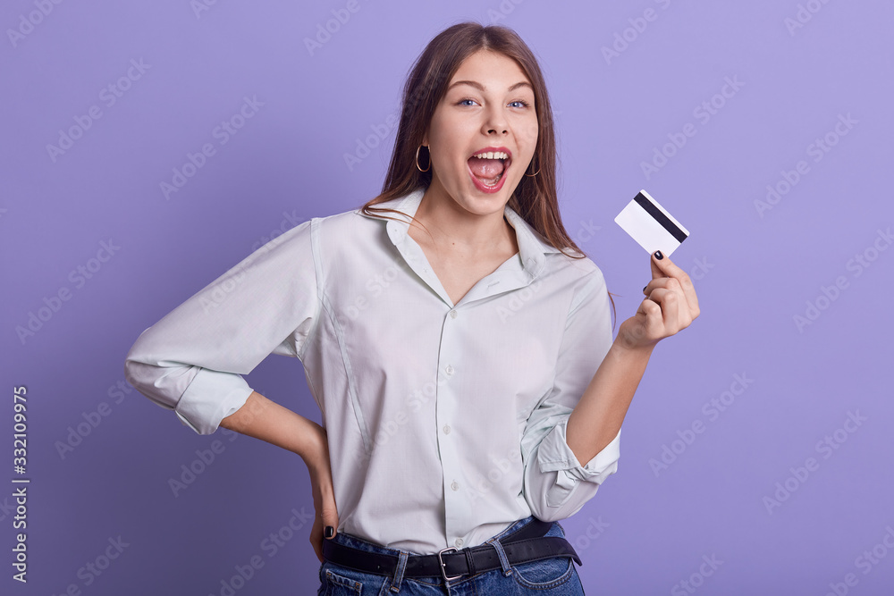 Indoor studio portrait of magnetic attractive young female posing isolated over lilac background, holding credit card, opening mouth, keeping one hand on waist, making credit payments successfully. Indoor studio portrait of magnetic attractive young female posing isolated over lilac background, holding credit card, opening mouth, keeping one hand on waist, making credit payments successfully.