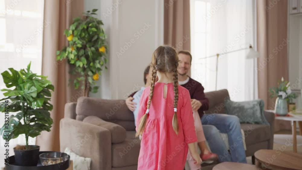 Follow slow motion shot of cute little girl with two braids walking to loving mother and father sitting on sofa at home. Middle aged parents enjoying talking with daughter sitting on mothers lap