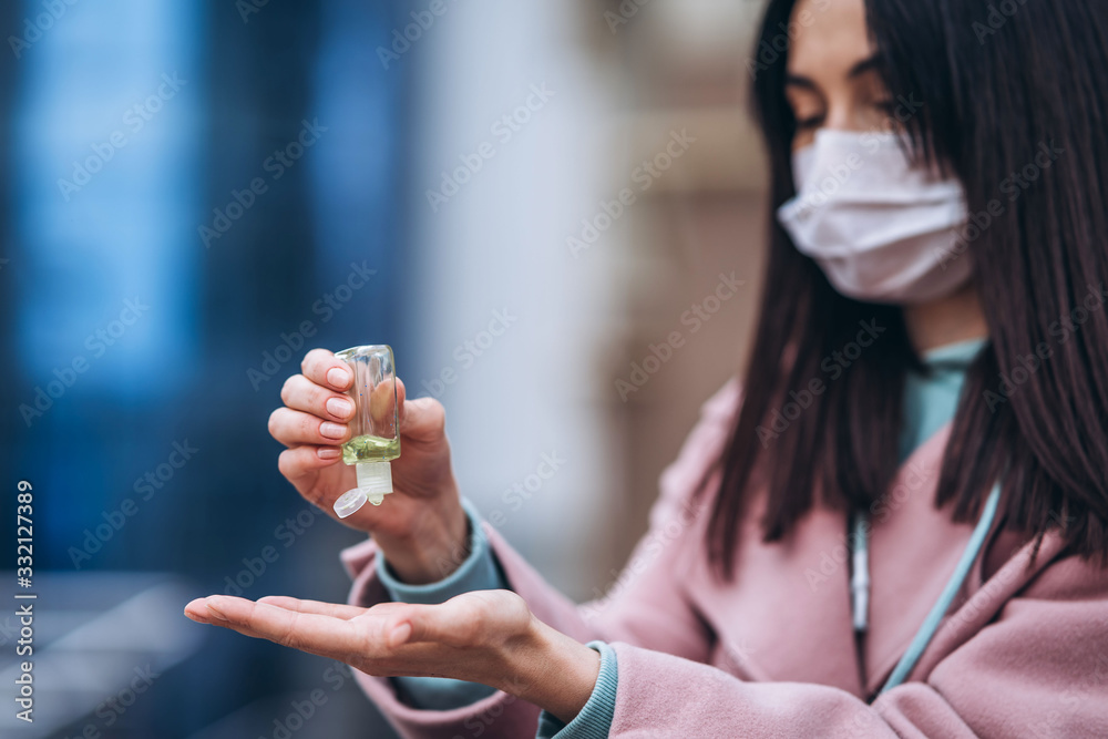 Fototapeta premium Close up hands of female in medical mask cleaning her hands with sanitizer outdoor in the city to prevent virus deseases. Coronavirus, COVID-19, epidemic, pandemic, quarantine concept.