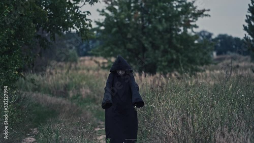 Dark figure in black robe with hood among nature. Scary nightmare walking on the natural background in summer. Mysterious ghost outdoors. photo