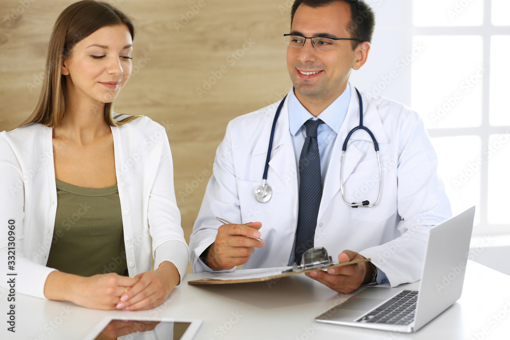 Doctor and patient discussing the results of a physical examination while sitting at a desk in a clinic. A male doctor using a clipboard to fill out a medical history of a young woman's medication Doctor and patient discussing the results of a physical examination while sitting at a desk in a clinic. A male doctor using a clipboard to fill out a medical history of a young woman's medication