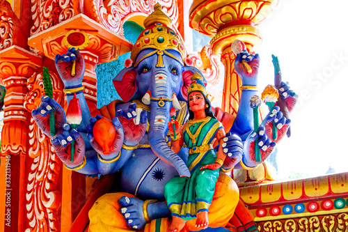 Photography Representation of Hindu gods and tourists climbing a colorful stairs leading to the Batu Caves in the background