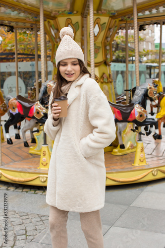 Street style. Caucasian girl posing in front of a carousel. Winter or autumn clothes. Pastel colors. Fun and smile. Merry-go-round.