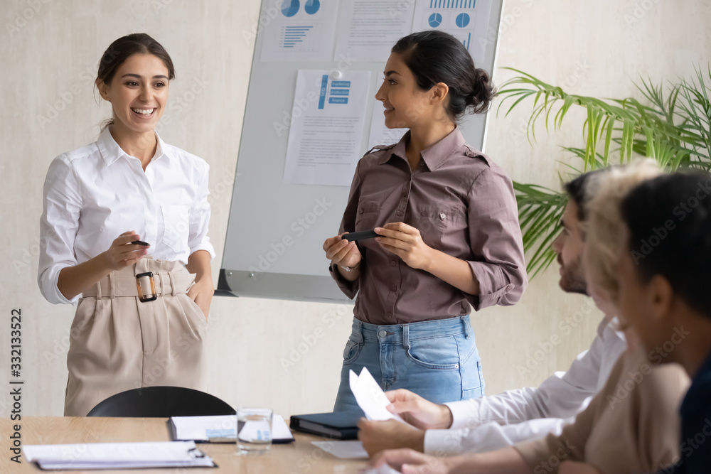 Happy female speakers talk make flip chart presentation to colleagues at meeting, smiling woman couch or tutors interact with coworkers at briefing, present business project on whiteboard in office Happy female speakers talk make flip chart presentation to colleagues at meeting, smiling woman couch or tutors interact with coworkers at briefing, present business project on whiteboard in office