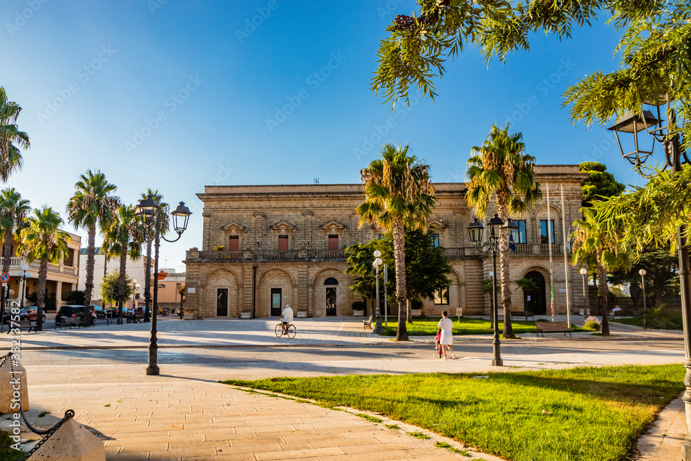 Acquarica del Capo, Presicce, Salento, Puglia, Italy. Town square with the seat of the municipality and the town hall. The ancient stone and brick building. Flower beds with the lawn and palm trees.