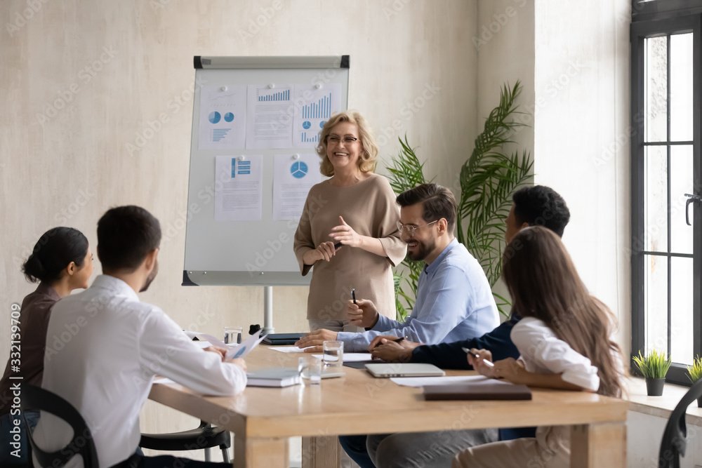 Smiling middle-aged female tutor or speaker talk make flip chart presentation to diverse colleagues at briefing, happy mature businesswoman joke presenting business project to coworkers at meeting Smiling middle-aged female tutor or speaker talk make flip chart presentation to diverse colleagues at briefing, happy mature businesswoman joke presenting business project to coworkers at meeting