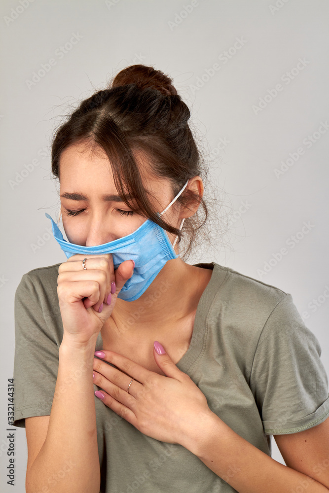 Young woman wearing protective mask and coughing. Protecting other from virus infection. Isolated on grey background.