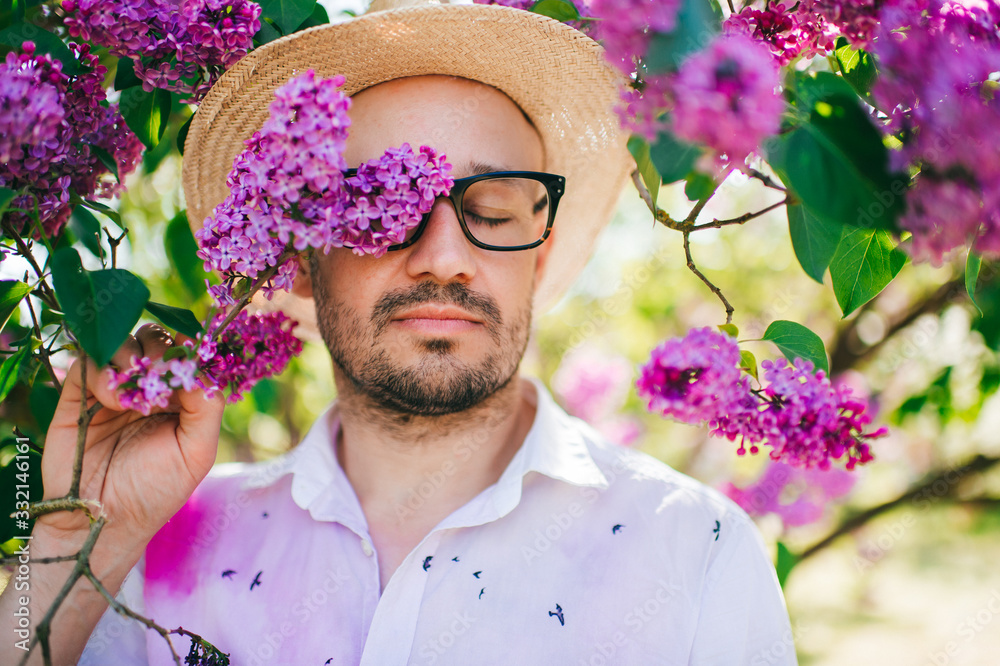 Obraz premium Portrait of beautiful man in a white hat with glasses stands in lilac garden and enjoys of flowers