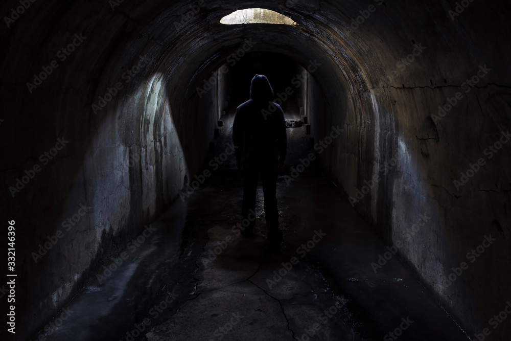 A figure of a man in a hood walking forward in a dark underground tunnel lit through a hatch in the ceiling. Fort Pospelova, Vladivostok.
