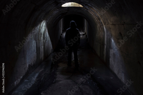 A figure of a man in a hood walking forward in a dark underground tunnel lit through a hatch in the ceiling. Fort Pospelova, Vladivostok. © Сергей Рамильцев