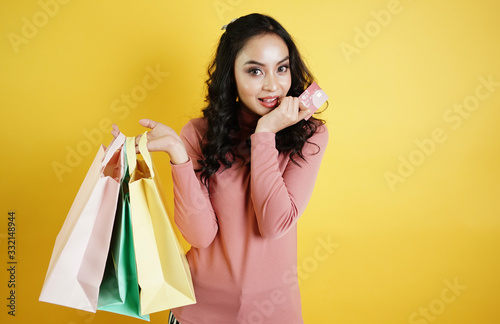Portrait of an excited beautiful Asian girl holding shopping bags and credit card isolated over Yellow background.