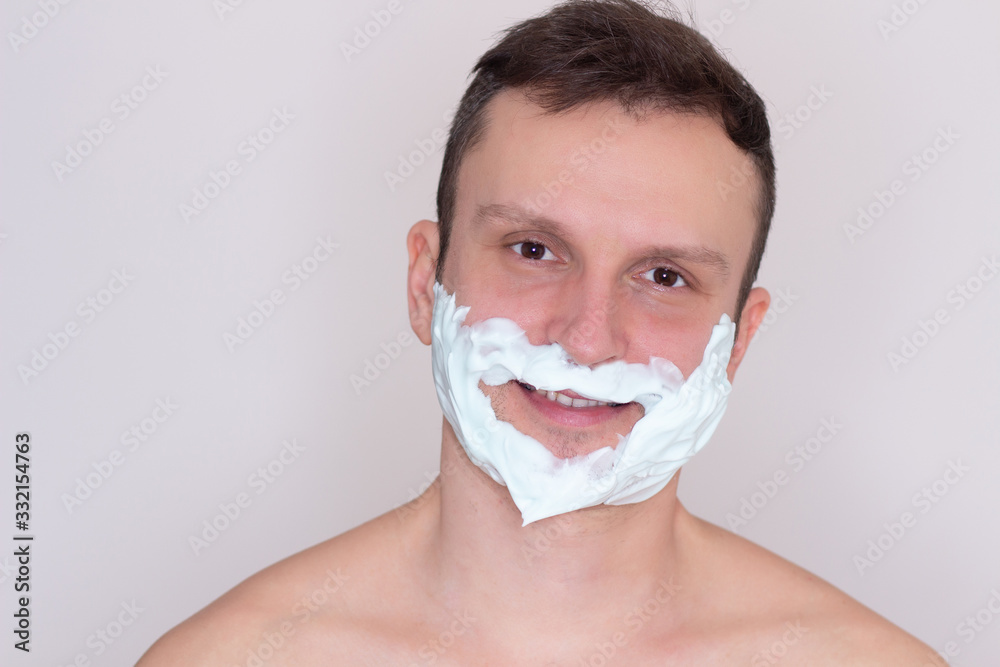 Portrait of young man shaving. He has white foam on beard. Guy is serious and concentrated. Isolated on white background.