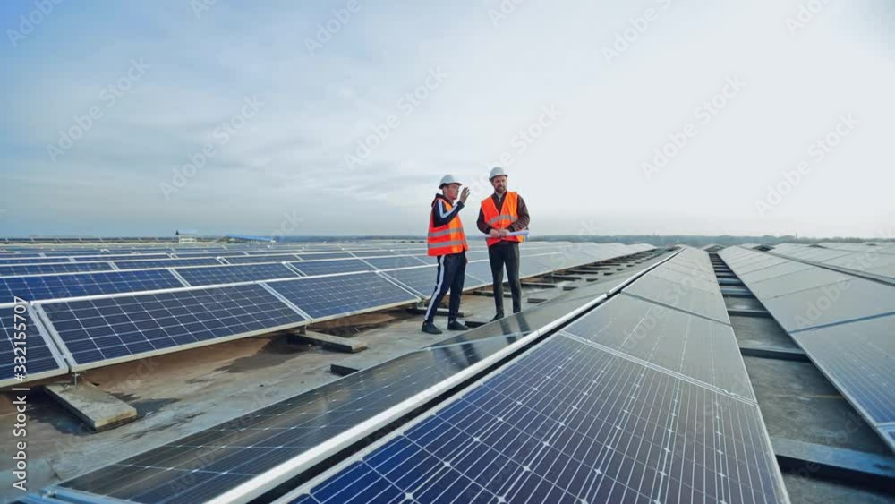 Modern solar farm on a roof under the sky. Workers in uniform and protective helmets discuss the solar panels installation.