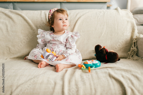 Little cute girl in beautiful dress sits on the couch and plays with her various toys