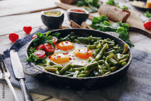 Pan of fresh fried eggs with tomatoes, green beans, bread, spices and herbs on rustic white wooden background