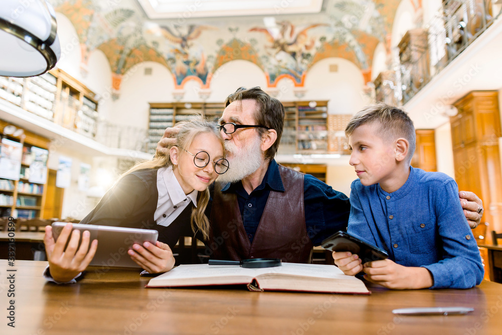 Elderly man teacher and his teen students, boy and girl, sitting at the table in library, reading book, using tablet and studying. Bearded man loves his students, is proud of them and kissing girl - obrazy, fototapety, plakaty 