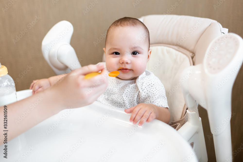 Baby eats in a highchair. Mom feeds the baby with pureed food. The first feeding of the baby. Baby eats in a highchair. Mom feeds the baby with pureed food. The first feeding of the baby.