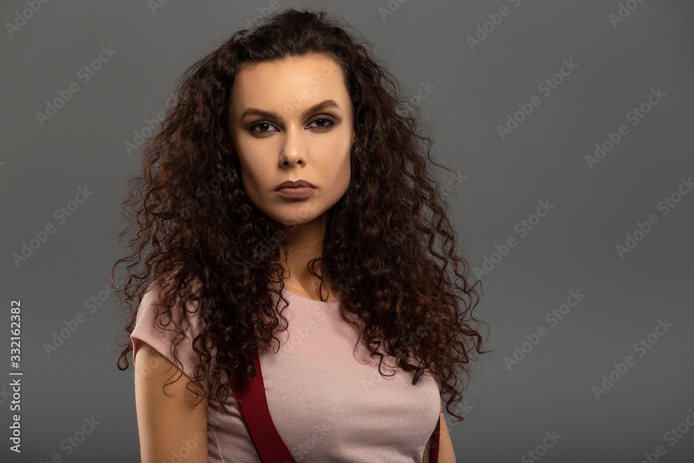 Young beautiful girl with curls on her head with a serious facial expression on a gray background. Portrait of a woman with a strong face with brown eyes in a pink T-shirt and red suspenders. Young beautiful girl with curls on her head with a serious facial expression on a gray background. Portrait of a woman with a strong face with brown eyes in a pink T-shirt and red suspenders.
