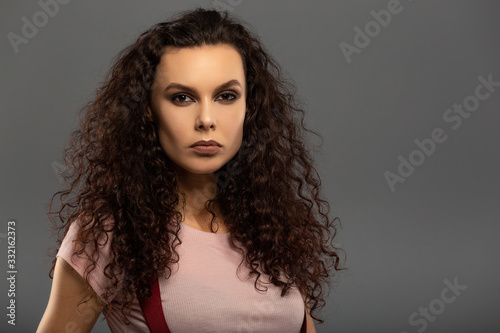 Young beautiful girl with curls on her head with a serious facial expression on a gray background. Portrait of a woman with a strong face with brown eyes in a pink T-shirt and red suspenders.