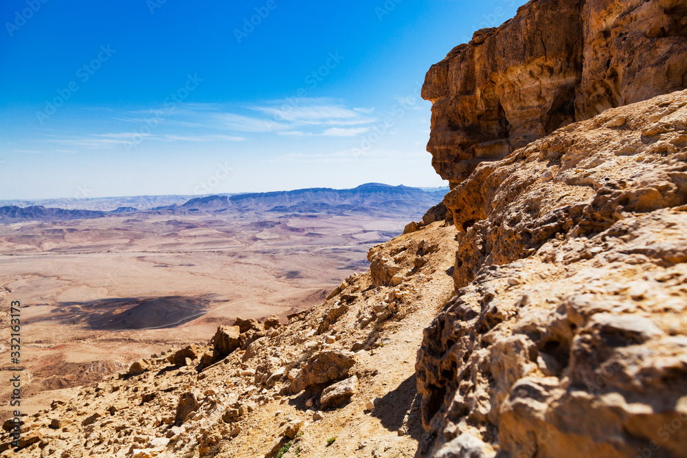 Beautiful view of the desert. A large rocky cliff and a desert valley on the horizon of which there are mountains. Blue sky and light white clouds. Monumentality and loneliness in a deserted place.