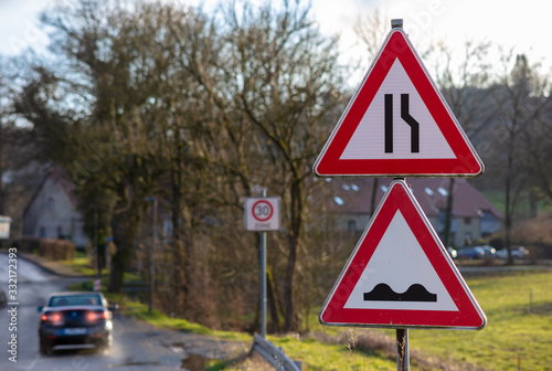 Two triangular red and white warning signs. The upper sign warns of a bottleneck. The lower sign warns of an uneven road or ruts. A car brakes in the background.