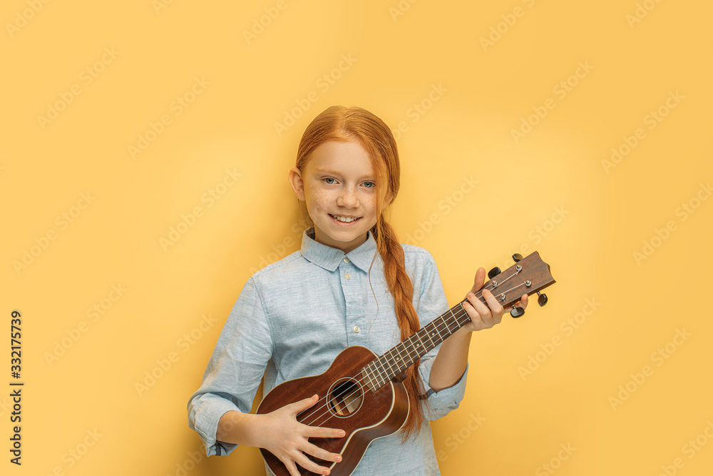 beautiful caucasian girl learns to play ukulele isolated over yellow background. attractive child with natural red hair hold small guitar, ukulele in hands and look at camera, smile, enjoy music