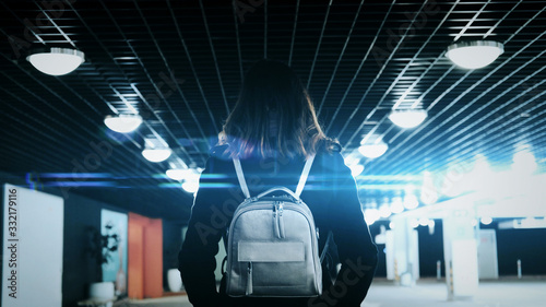 A girl in a black coat and with a backpack walks through an underground parking lot. An anamorphic glare, cinematic picture. The woman's view from the back. photo