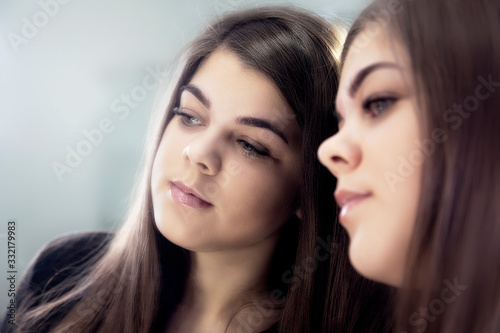 Portrait of a young beautiful girl near the mirror. Girl and her reflection in the mirror. The girl looks at the camera through the mirror. Selective focus.