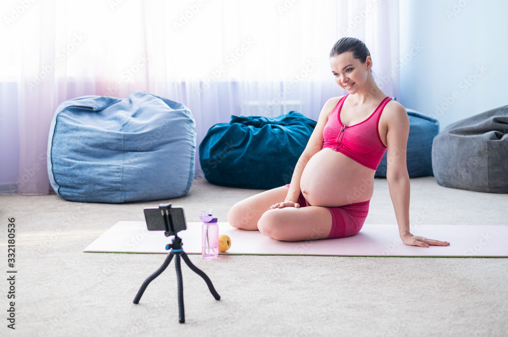 Fototapeta premium Online lessons for pregnant women. European woman shoots a video fitness lesson on the phone. Waiting for a miracle. Yoga for expectant mothers.