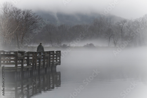 Pensive Older Man Stands Alone By a Foggy Mountain Lake in Winter photo