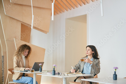 Wide angle portrait of two young women in business casual working or studying sitting at separate tables in indoor cafe, copy space