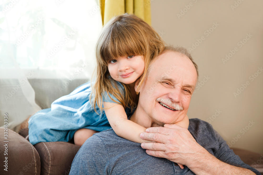 Happy grandpa and child granddaughter hugging and  looking at camera and smiling while resting together at home. generations family