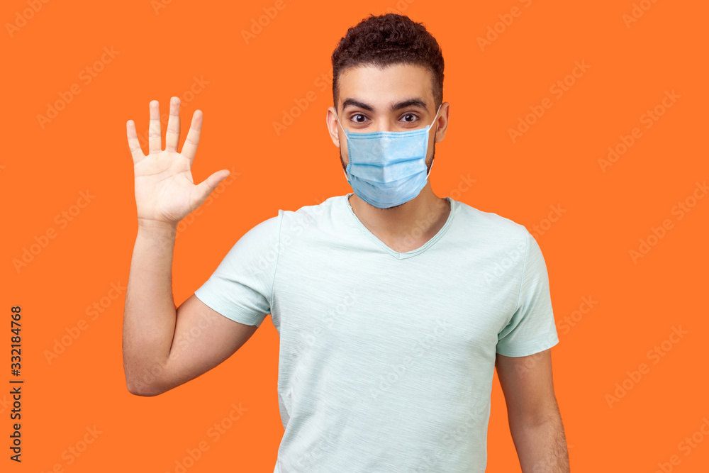 Hello! Portrait of friendly glad handsome man with medical mask in casual white t-shirt showing hi gesture with waving hand and smiling sincerely. indoor studio shot isolated on orange background Hello! Portrait of friendly glad handsome man with medical mask in casual white t-shirt showing hi gesture with waving hand and smiling sincerely. indoor studio shot isolated on orange background