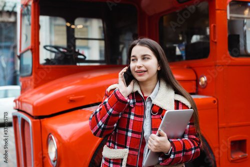 beautiful girl in checkered jacket with laptop talking on smartphone near red bus on city street © MarHo