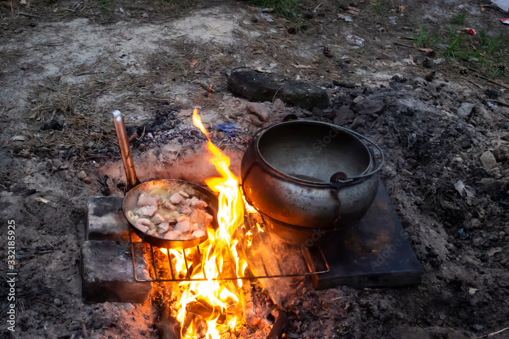 Field kitchen in the forest. Water boils and cooking food. Boiling pot and frying pan above the fire, tourist meal cooking on fire Field kitchen in the forest. Water boils and cooking food. Boiling pot and frying pan above the fire, tourist meal cooking on fire