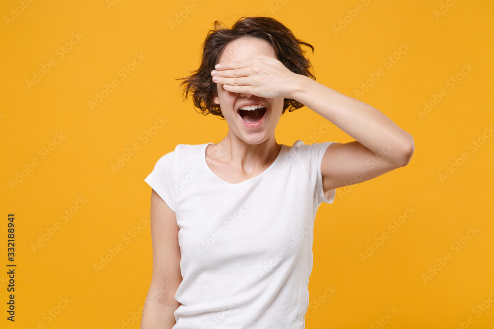 Cheerful young brunette woman girl in white t-shirt posing isolated on yellow orange background studio portrait. People emotions lifestyle concept. Mock up copy space. Covering eyes with hand, hiding. Cheerful young brunette woman girl in white t-shirt posing isolated on yellow orange background studio portrait. People emotions lifestyle concept. Mock up copy space. Covering eyes with hand, hiding.