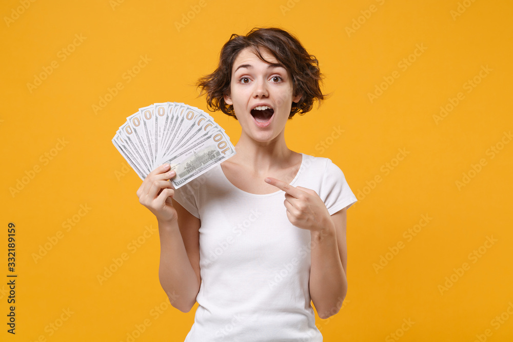 Shocked young brunette woman in white t-shirt posing isolated on yellow orange background. People lifestyle concept. Mock up copy space. Pointing index finger on fan of cash money in dollar banknotes. Shocked young brunette woman in white t-shirt posing isolated on yellow orange background. People lifestyle concept. Mock up copy space. Pointing index finger on fan of cash money in dollar banknotes.