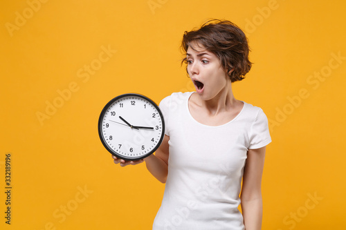 Shocked young brunette woman girl in white t-shirt posing isolated on yellow orange wall background studio portrait. People sincere emotions lifestyle concept. Mock up copy space. Hold in hand clock.