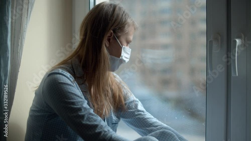 Young beautiful woman in home clothes and a protective mask sitting on the windowsill and looking out the window. Quarantine during the coronavirus epidemic. COVID-19 pandemic photo