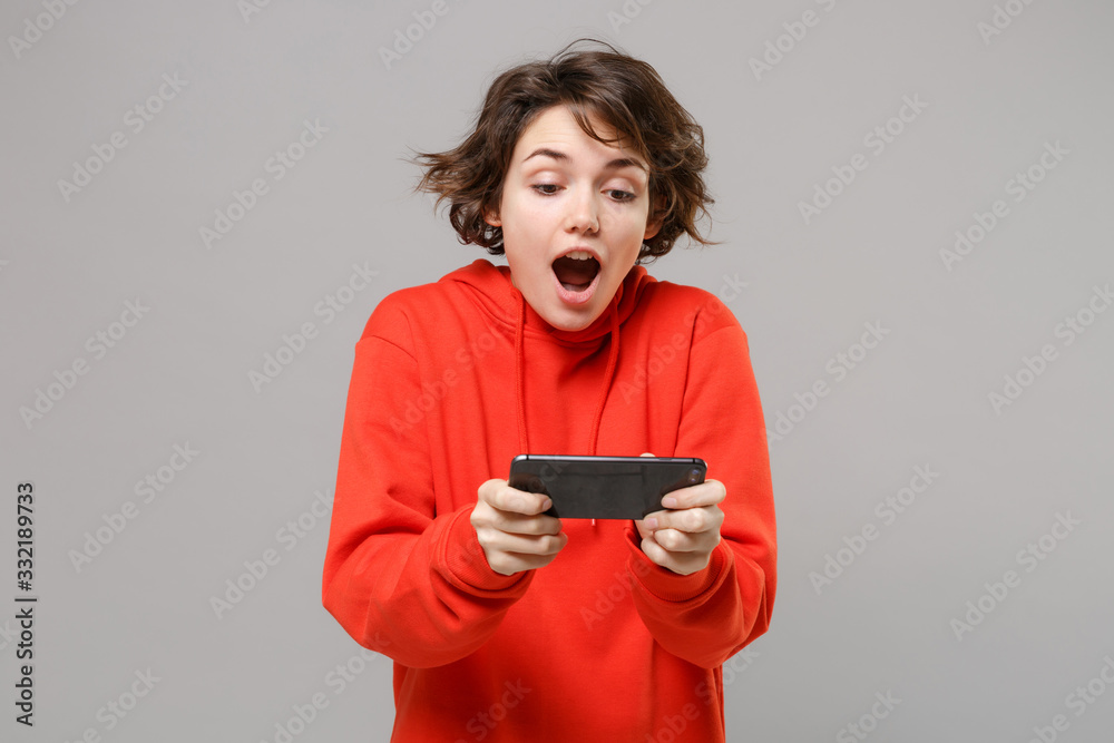Excited young brunette woman girl in casual red hoodie posing isolated on grey background studio portrait. People sincere emotions lifestyle concept. Mock up copy space. Play game with mobile phone. Excited young brunette woman girl in casual red hoodie posing isolated on grey background studio portrait. People sincere emotions lifestyle concept. Mock up copy space. Play game with mobile phone.