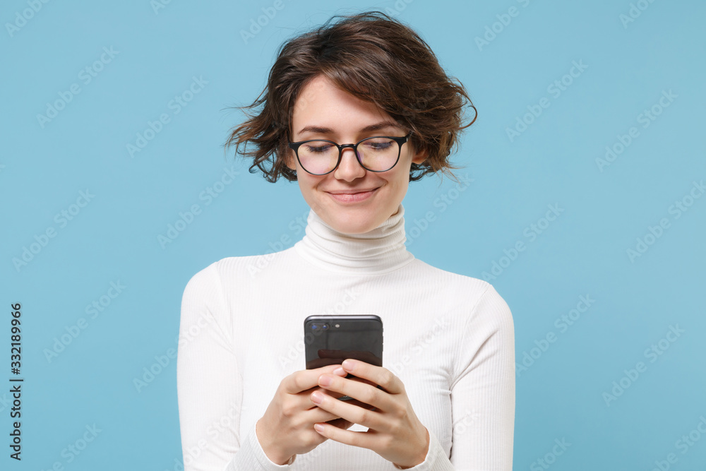 Smiling young brunette woman girl in casual white clothes, eyeglasses posing isolated on pastel blue background. People lifestyle concept. Mock up copy space. Using mobile phone, typing sms message. Smiling young brunette woman girl in casual white clothes, eyeglasses posing isolated on pastel blue background. People lifestyle concept. Mock up copy space. Using mobile phone, typing sms message.