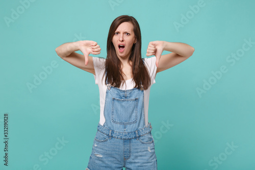 Irritated shocked young brunette woman girl in casual denim clothes posing isolated on blue turquoise background studio portrait. People lifestyle concept. Mock up copy space. Showing thumbs down.
