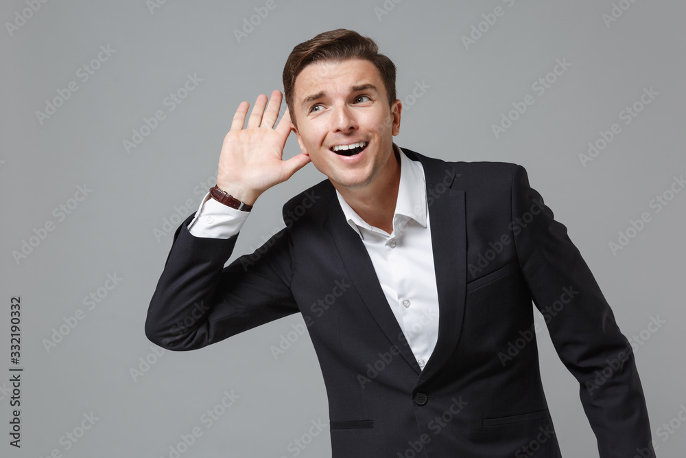 Curious young business man in classic black suit shirt posing isolated on grey background in studio. Achievement career wealth business concept. Mock up copy space. Try to hear you with hand near ear. Curious young business man in classic black suit shirt posing isolated on grey background in studio. Achievement career wealth business concept. Mock up copy space. Try to hear you with hand near ear.