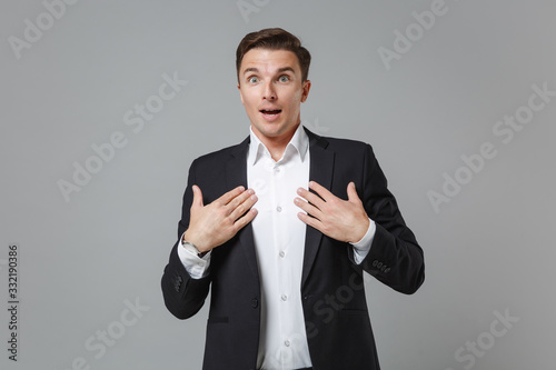 Surprised young business man in classic black suit shirt posing isolated on grey background studio portrait. Achievement career wealth business concept. Mock up copy space. Keeping hands on chest.