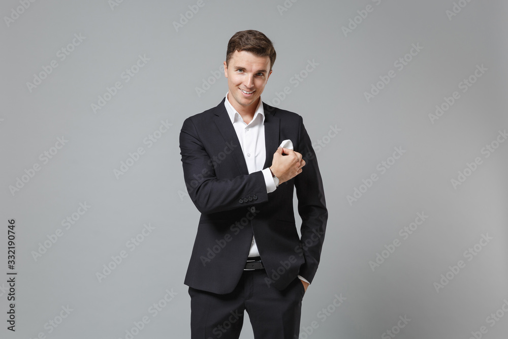 Smiling young business man in classic black suit shirt posing isolated on grey background. Achievement career wealth business concept. Mock up copy space. Straightening handkerchief in chest pocket. Smiling young business man in classic black suit shirt posing isolated on grey background. Achievement career wealth business concept. Mock up copy space. Straightening handkerchief in chest pocket.